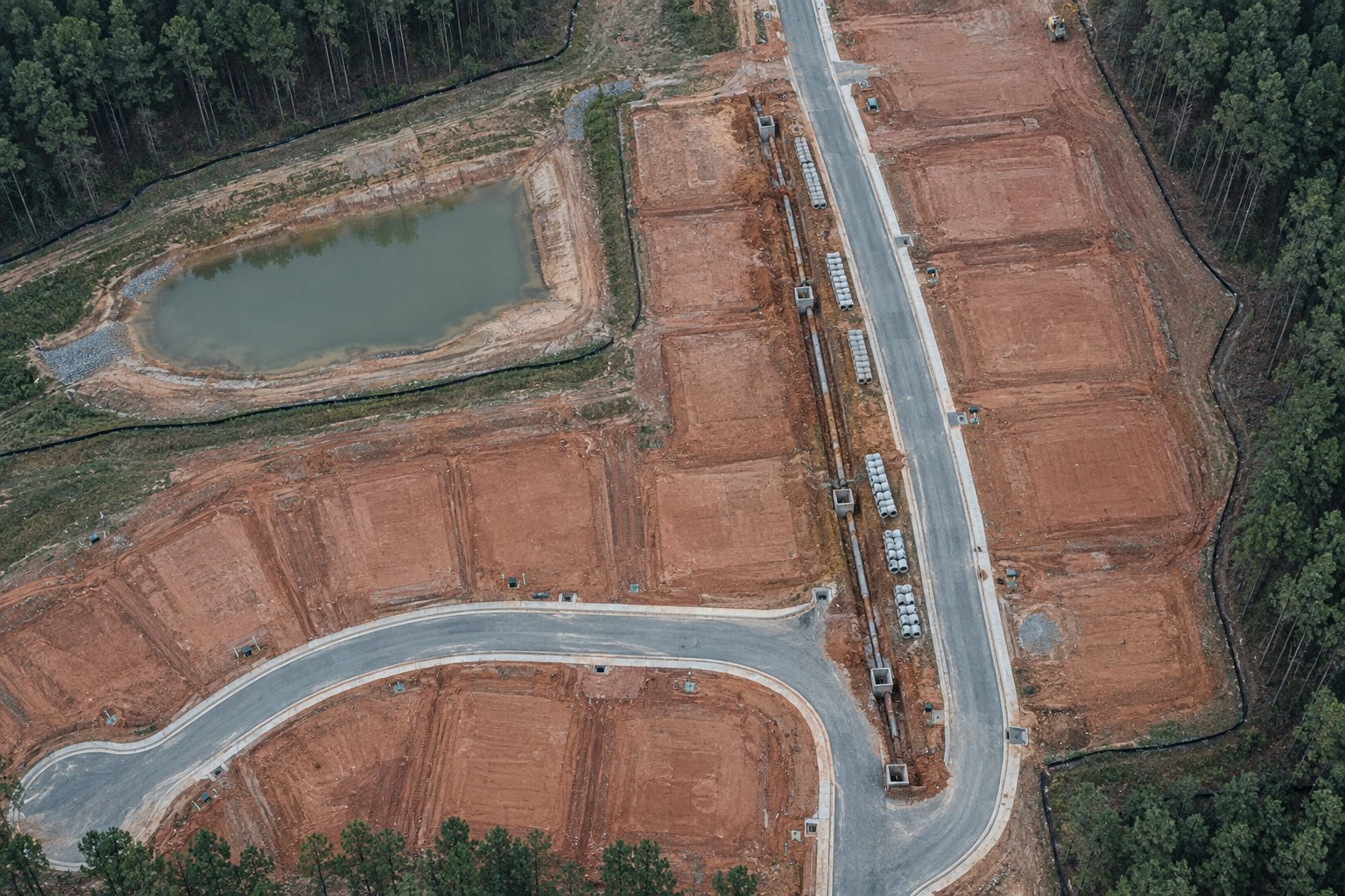Aerial of a subdivision under construction: graded pads, a new street, a storm pipe trench, and a pond